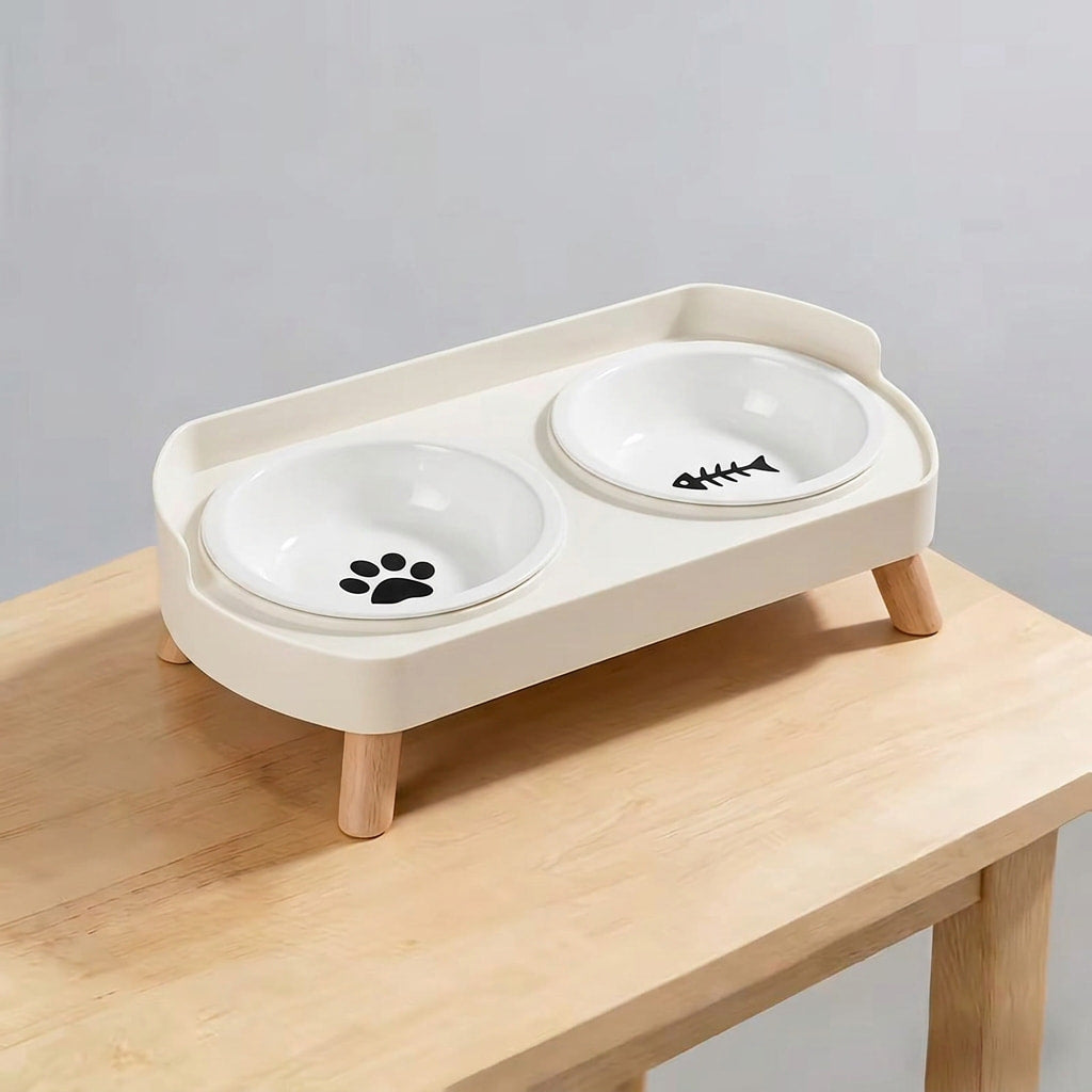 A two-bowl elevated pet feeder and waterer with a cream-colored plastic tray and wooden legs, shown on a light wood table against a neutral gray background.
