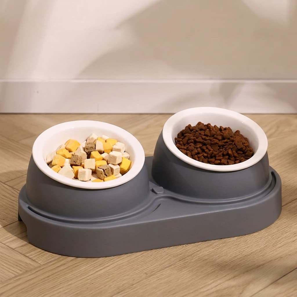 A dual elevated cat feeder bowl set in grey with white bowls, one filled with mixed wet pet food and the other with dry kibble, placed on a light wood floor against a beige wall.