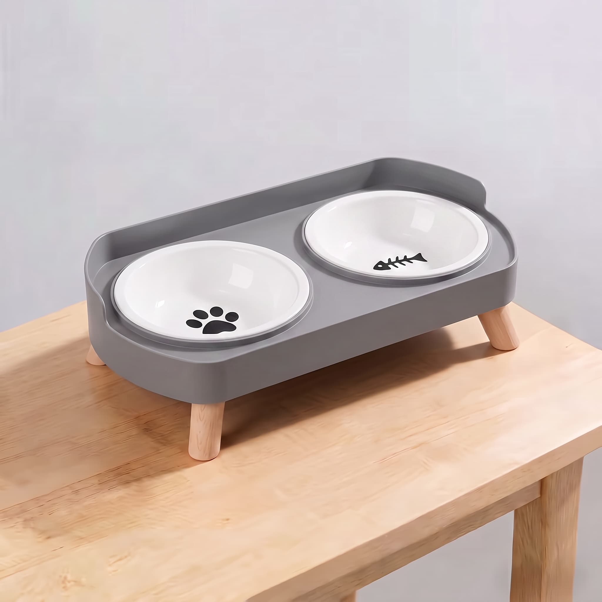 A grey elevated double pet feeder with two white ceramic bowls on wooden legs, shown on a light wood table against a plain light grey wall. One bowl has a black paw print and the other has a fish skeleton graphic.