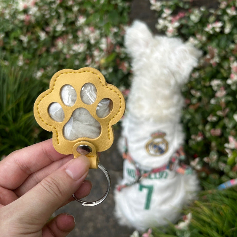 Yellow paw-shaped keychain held by a hand with a white dog in the background.
