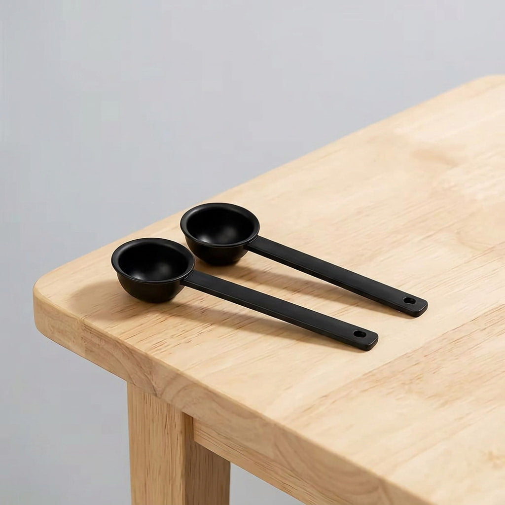 Two matte black silicone measuring cups with long handles, resting on a light-colored wooden table against a neutral gray background.