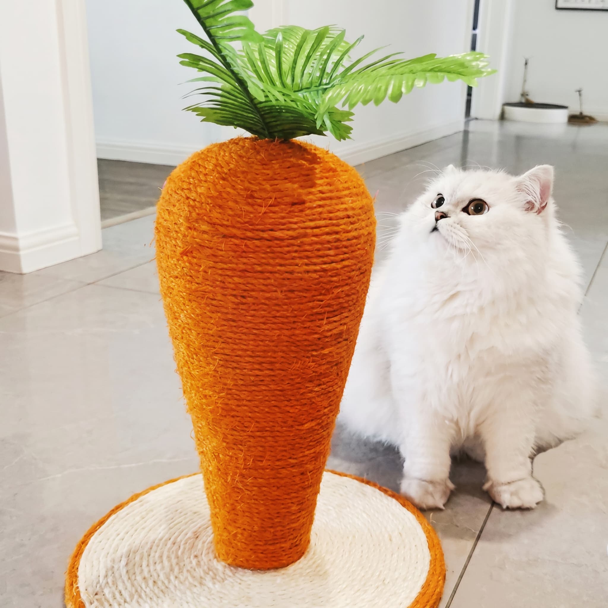 Cat toy shaped like a carrot with a green top, resembling a plant, next to a white cat indoors.