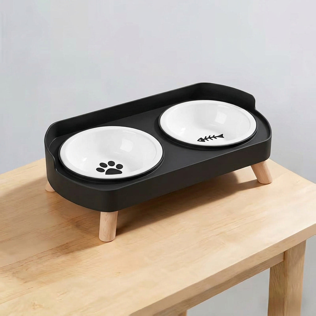 A two-bowl elevated pet feeder and waterer stand in matte black with two white ceramic bowls, shown resting on a light wood table against a neutral gray background.