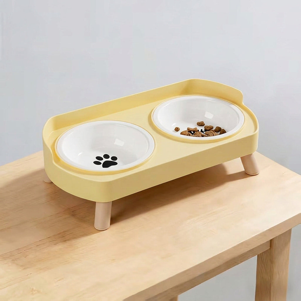 A yellow elevated double pet feeder with two white bowls, one with a black paw print and the other filled with kibble, shown on a light wood table against a neutral gray background.