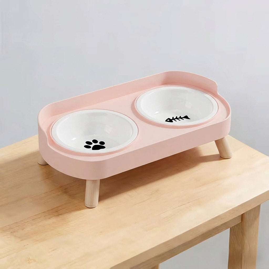 A pink elevated double pet feeder and water bowl stand with white ceramic bowls, shown on a light wood table against a plain light gray wall.
