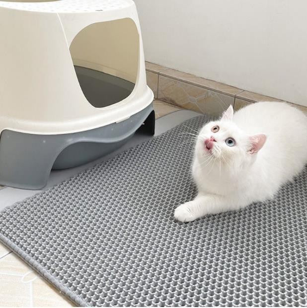 White cat on a grey mat next to a cat litter box in a bathroom.