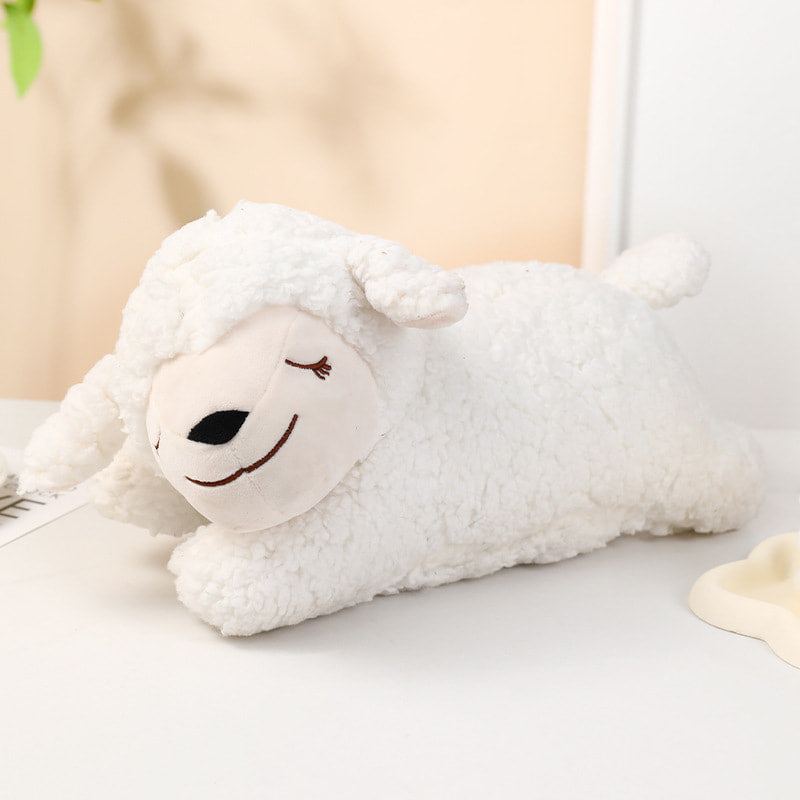 A white plush sheep toy with a soft fuzzy texture, lying down on a light-colored surface. The sheep has a cream-colored face with embroidered black eyes and a brown stitched smile, set against a softly blurred background.