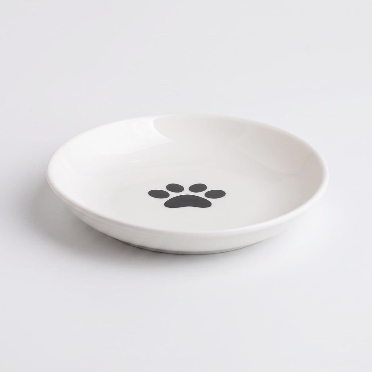 A white ceramic shallow bowl with a black paw print design in the center, photographed on a clean white background.