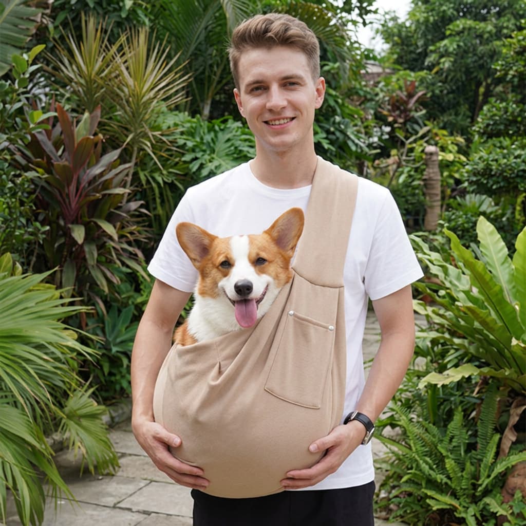 A man wearing a white t-shirt and black pants carries a corgi dog in a beige pet carrier backpack, set against a lush green garden background.