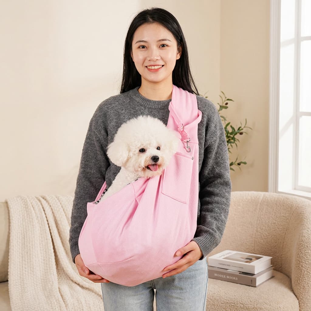A woman wearing a grey sweater and light blue jeans holds a small white dog in a pink pet carrier sling. The scene is set in a cozy, softly lit living room with a beige sofa, a blanket, and a window in the background.