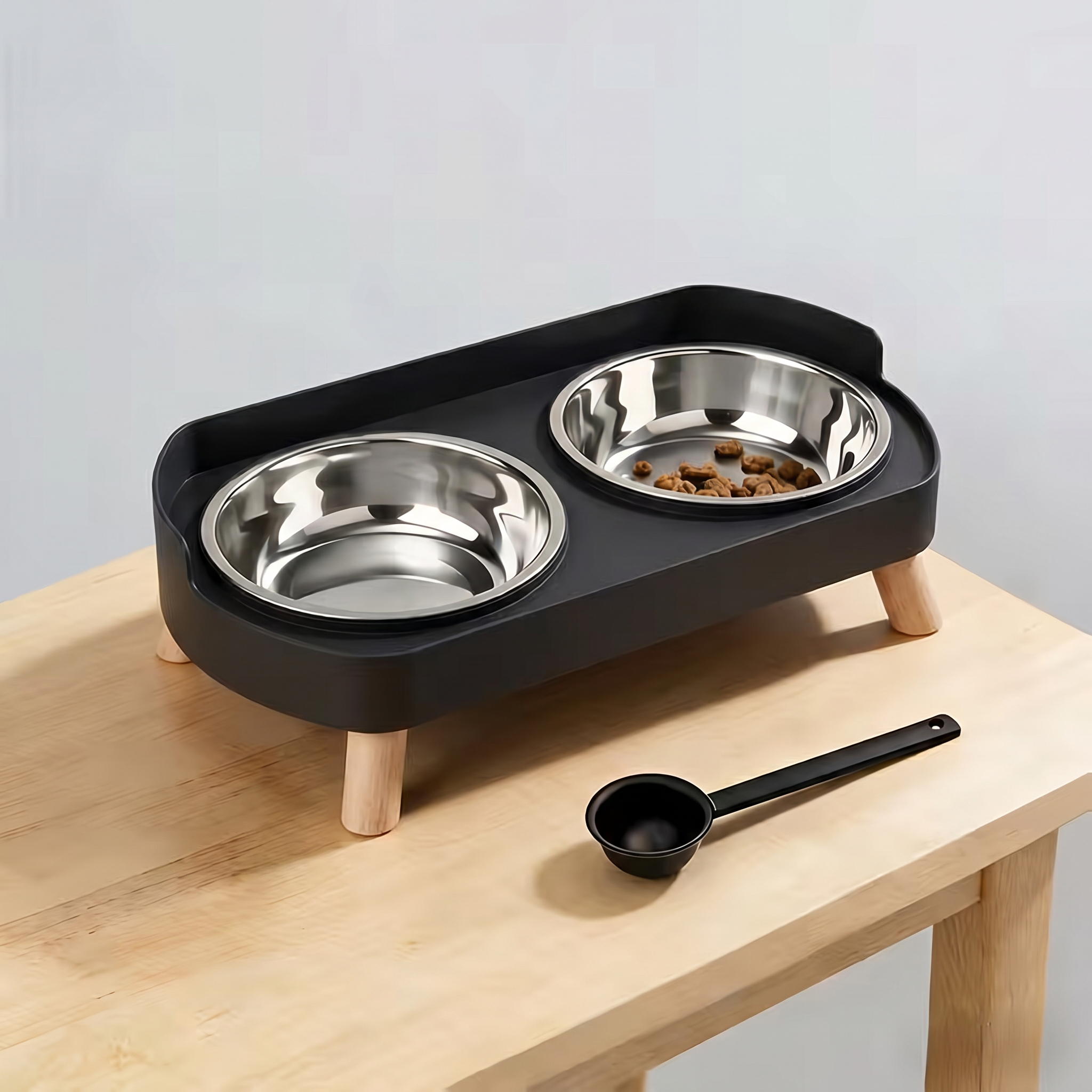 Pet feeding station with two stainless steel bowls on a black elevated platform, accompanied by a black scoop on a wooden surface.