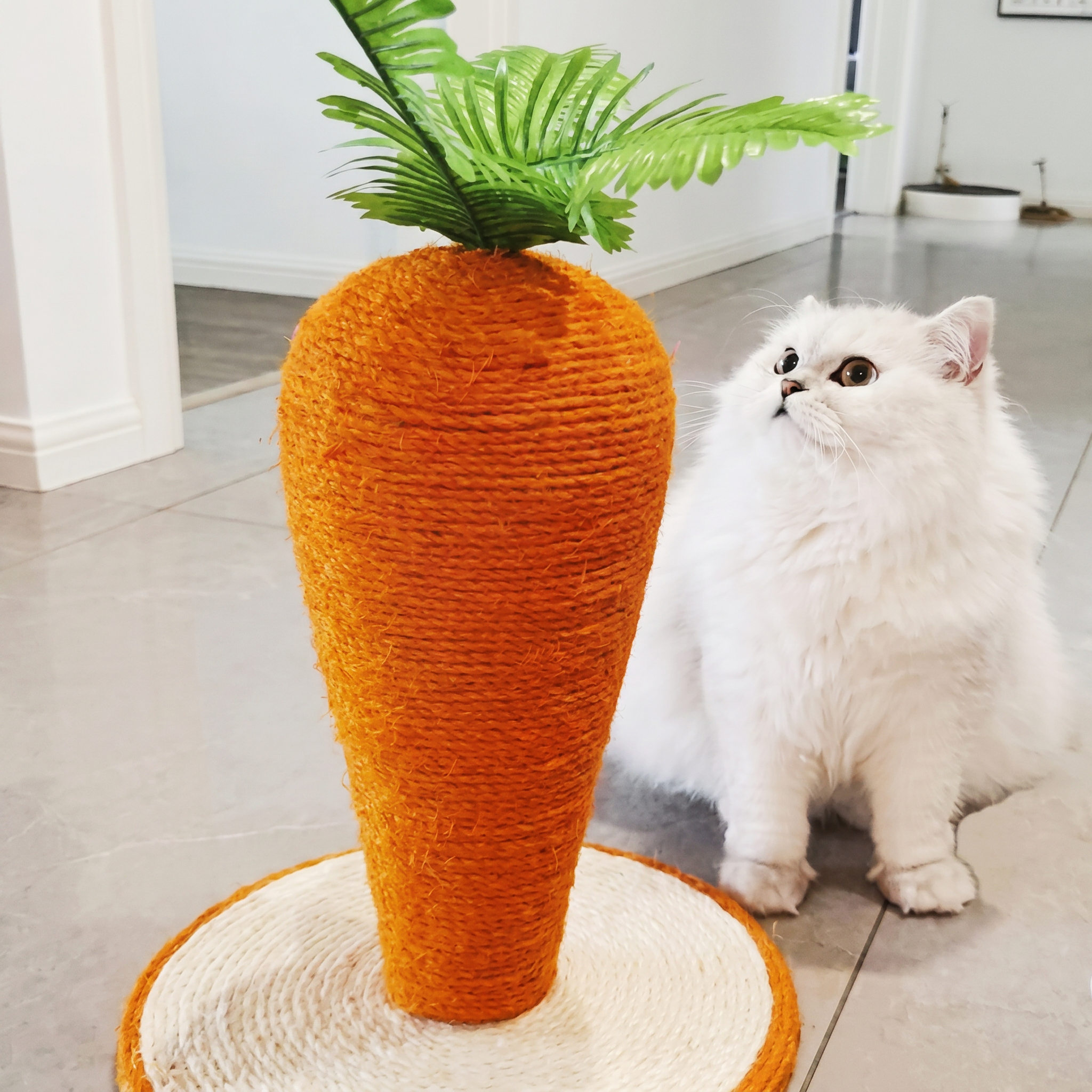 Cat toy shaped like a carrot with a green top, resembling a plant, next to a white cat indoors.